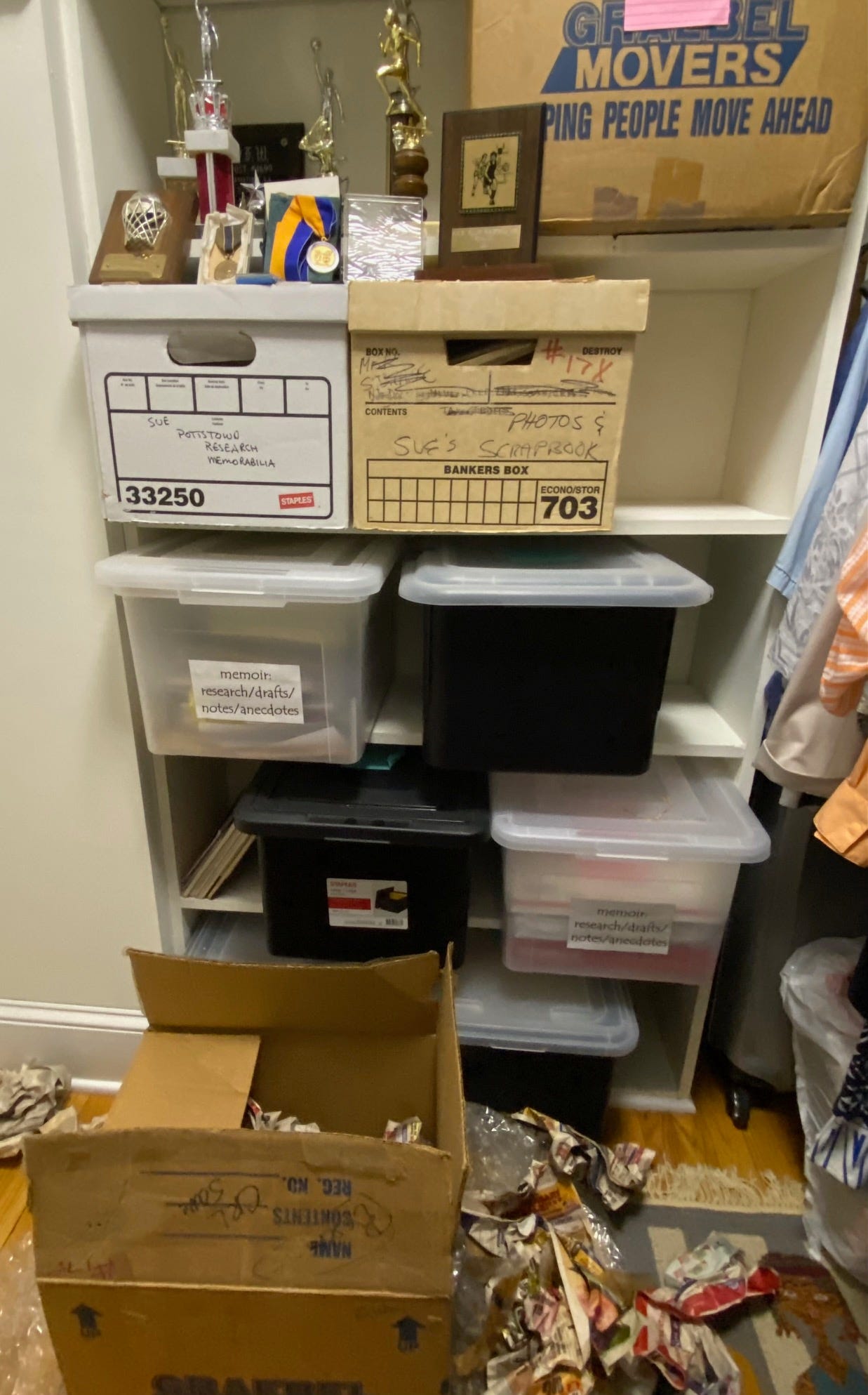 Photo of plastic and cardboard boxes on shelves in a clothes closet. On the top shelf are various trophies and medals. On the floor is an opened box with crumpled newspapers and bubble wrap in and around it.