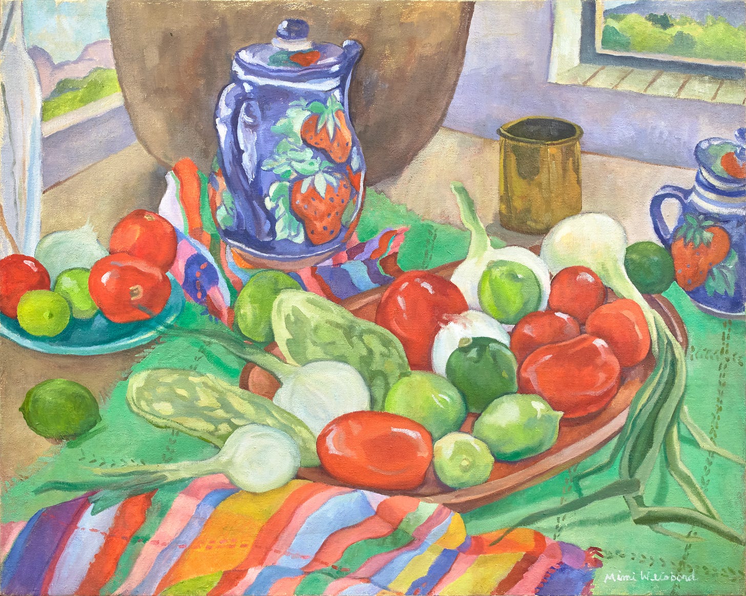 A table holding bowls tumbling with fresh fruit and vegetables with Mexican textiles in the same colors and pottery painted with images of strawberries. Behind are windows overlooking a green landscape with blue sky and hills.