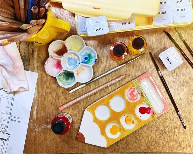 a photograph of a disorganized studio desk with many art materials. a pencil shaped paint palette is surrounded by paintbrushes, pencils, inks, paints, and more.