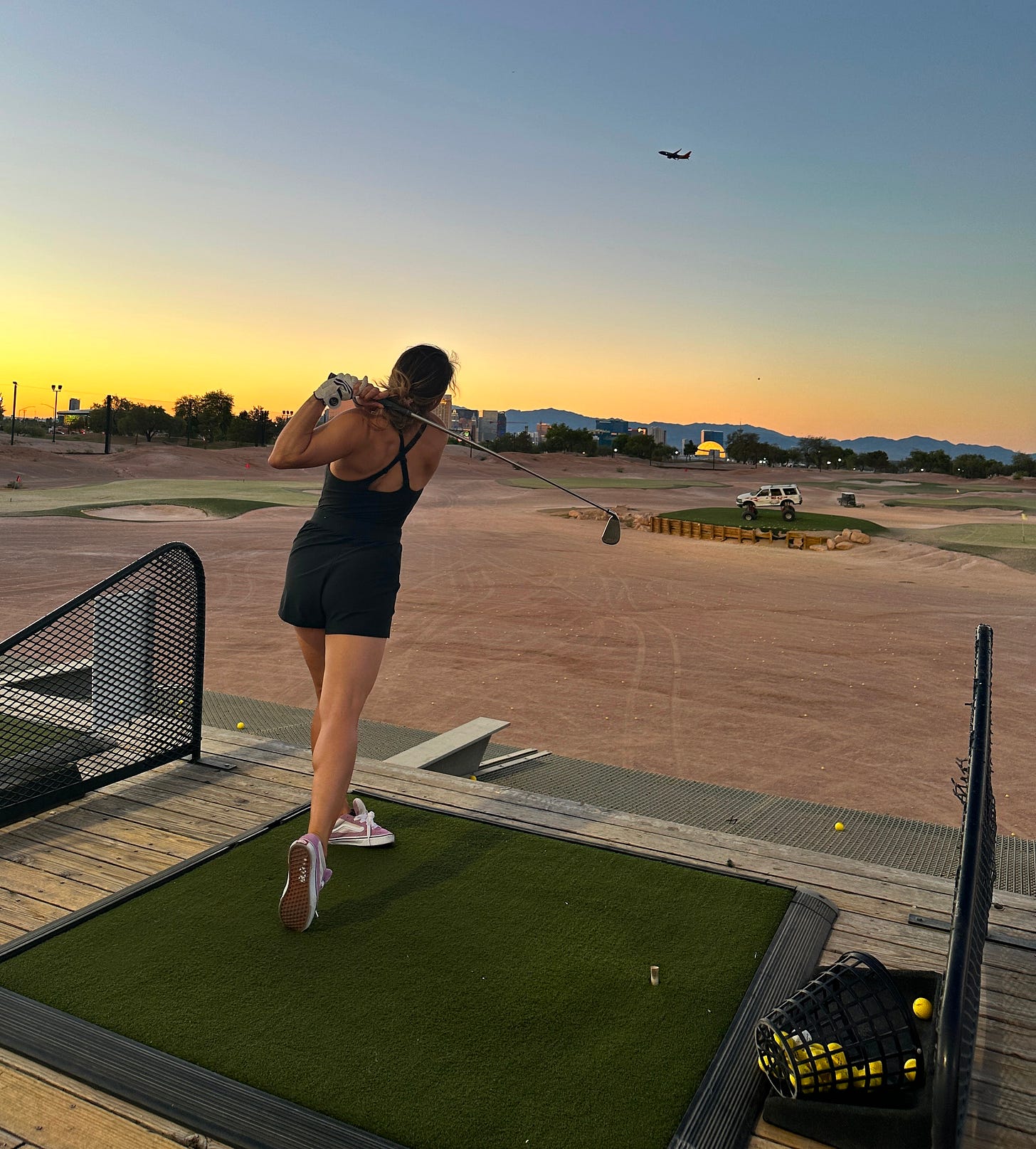 Pamela Maldonado finishing a golf swing at sunset, facing a desert driving range with mountains and an airplane in the distance in Las Vegas.