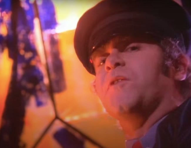 Elton John en chemise brune et casquette noire, menton en avant et regard sombre, dans le clip "Fascist Faces"