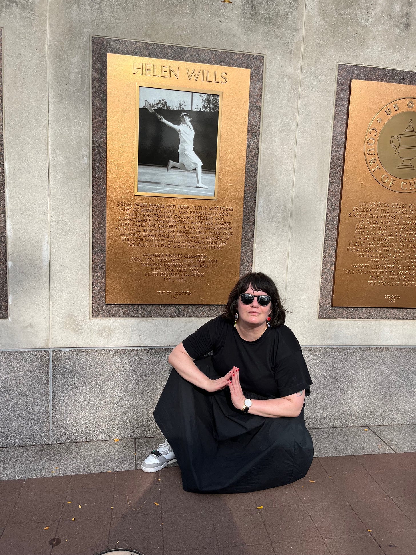 Me in a black dress and Chlorophyll Air Trainers kneeling in front of a plaque of tennis legend, Helen Wills, at the US Open.