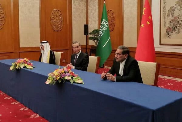 Officials from Saudi Arabia, China and Iran sit in a line at a blue table in a wood paneled room with their countries' flags behind them.
