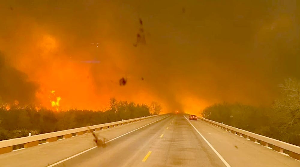 Soot collects on a windshield of a vehicle on a two lane highway facing a glowing orange sky and heavy smoke.