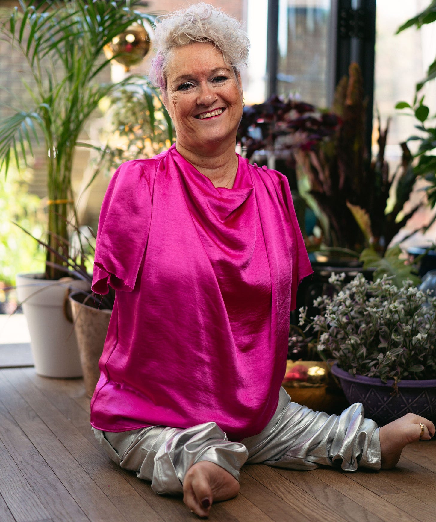 A middle aged disabled woman with no arms is sitting on the floor, surrounded by plants, wearing shimmering metallic clothes and smiling widely.