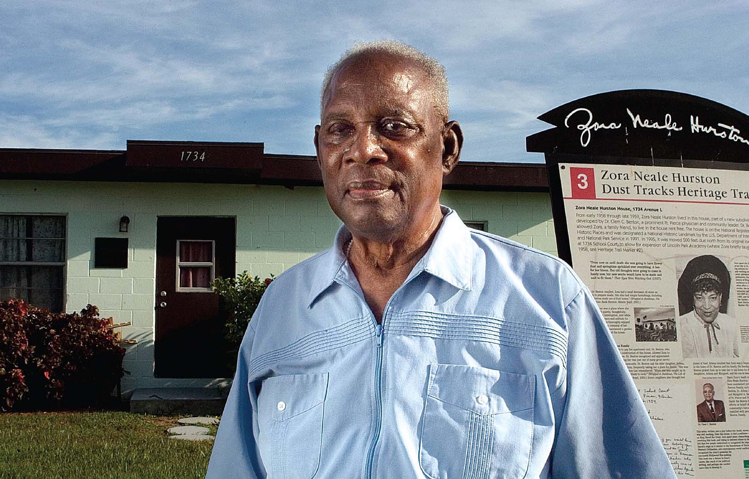 photo of an older black man in a blue collared shirt standing in front of a boxy green house and a sign that identifies the house as Zora Neale Hurston's