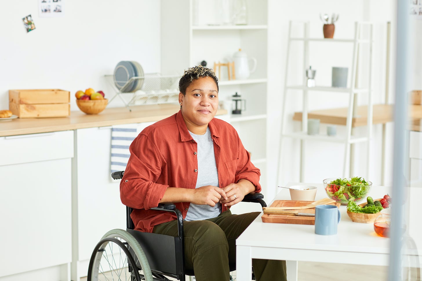 A person of color sits in a manual wheelchair in a kitchen.