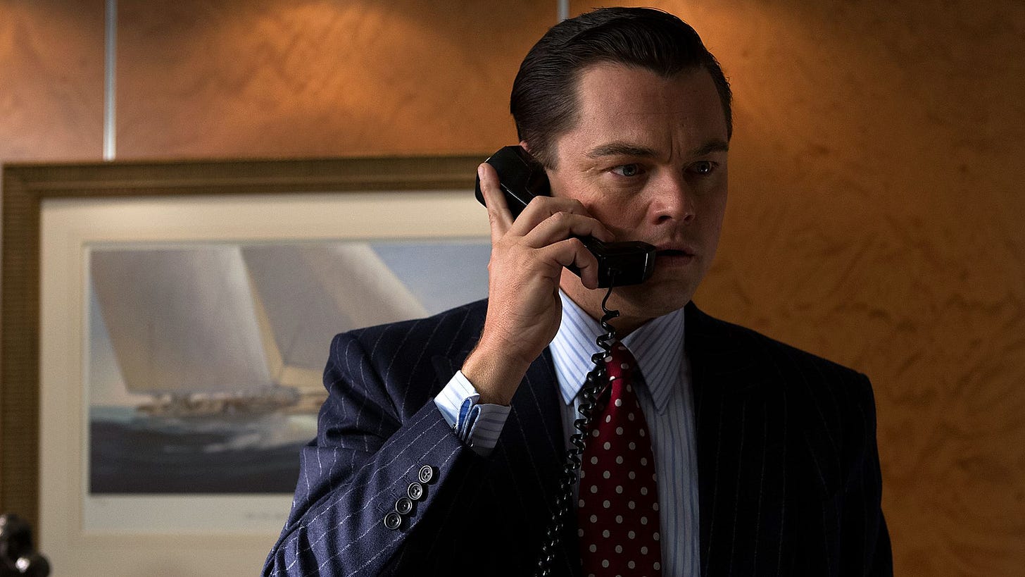 Leonardo di Caprio holds a phone, a shocked expression on his face, a very sharp pin-striped suit and spotted tie, a painting of a racing yacht under sail behind him, in the film The Wolf of Wall Street