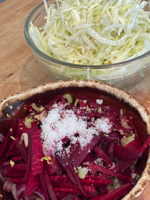Foreground a bowl with slices of beets, celery and red onion with a dash of salt on top. In the background a bowl with shredded fennel and cabbage. 