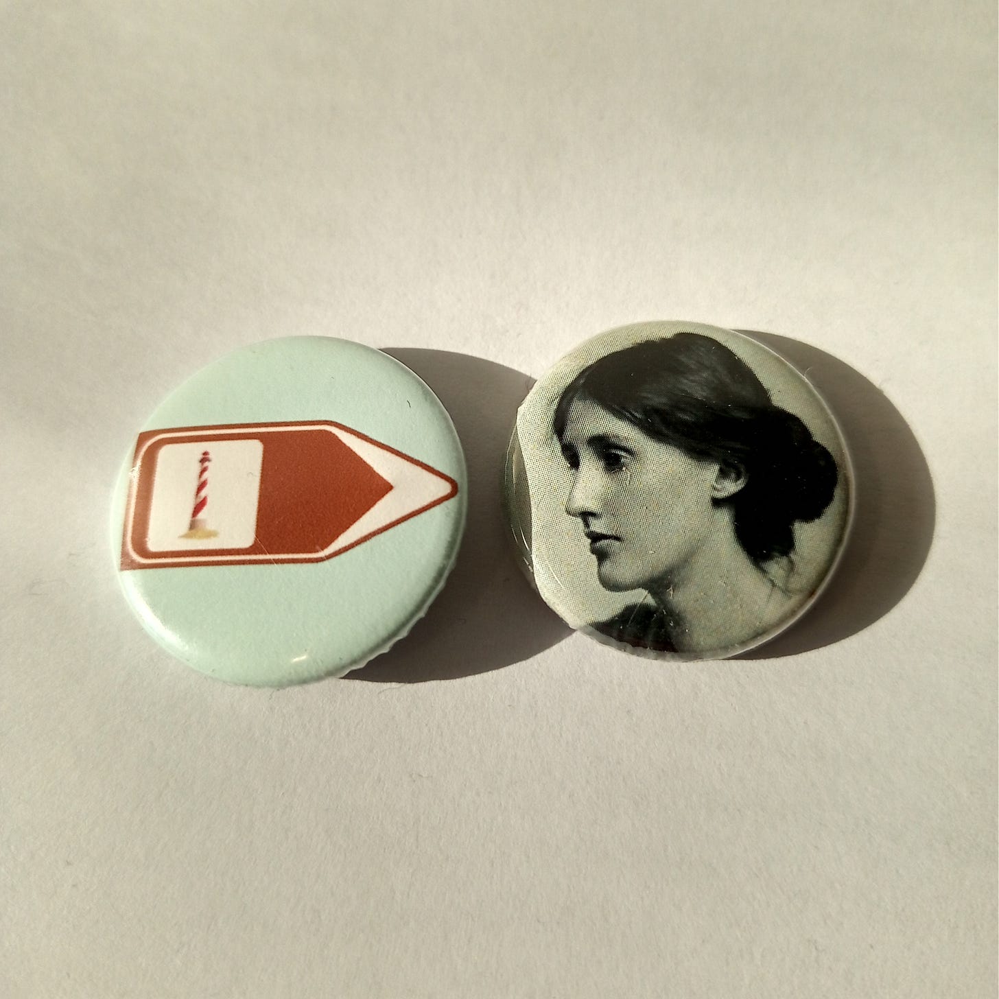 Two button badges in sunlight, one with a tourist sign pointing towards a lighthouse, one with a photograph of Virginia Woolf in profile