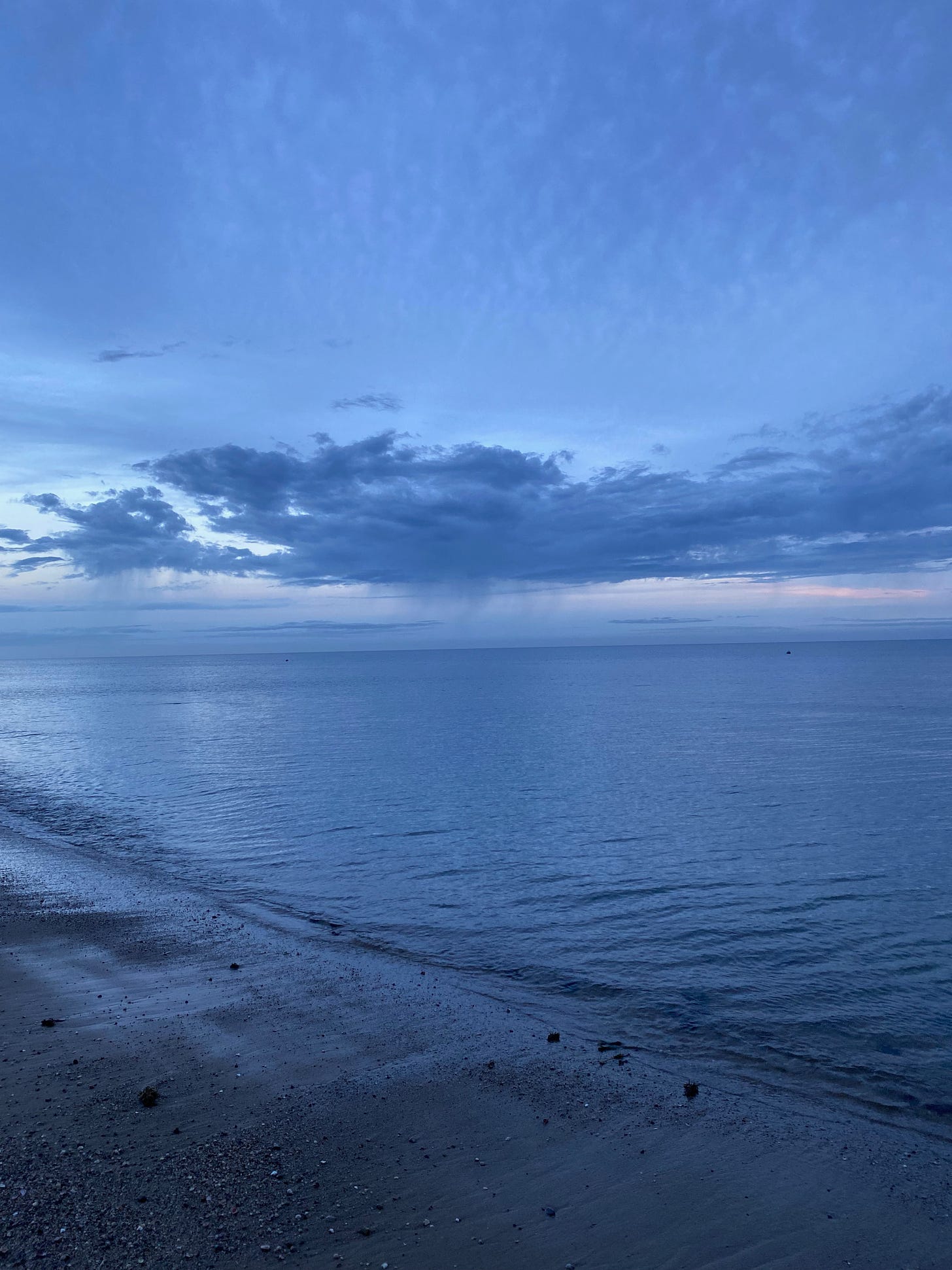 The beach at sunrise. The ocean is still and blue, and the sky is deep blue, with dark clouds, and silver and pink streaks along the horizon.