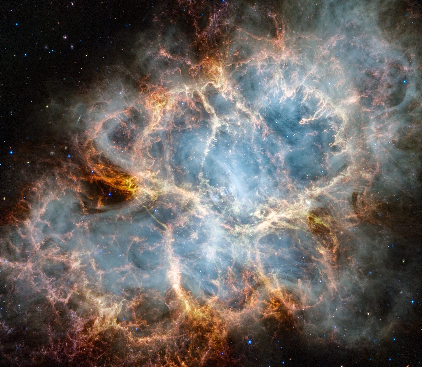 Webb image of the Crab Nebula. An oval nebula with complex structure against a black background. On the nebula’s exterior, particularly at the top left and bottom left, lie curtains of glowing red and orange fluffy material. Its interior shell shows large-scale loops of mottled filaments of yellow-white and green, studded with clumps and knots. Translucent thin ribbons of smoky white lie within the remnant’s interior, brightest toward its center. The white material follows different directions throughout, including sometimes sharply curving away from certain regions within the remnant. A faint, wispy ring of white material encircles the very center of the nebula. Around and within the supernova remnant are many points of blue, red, and yellow light.