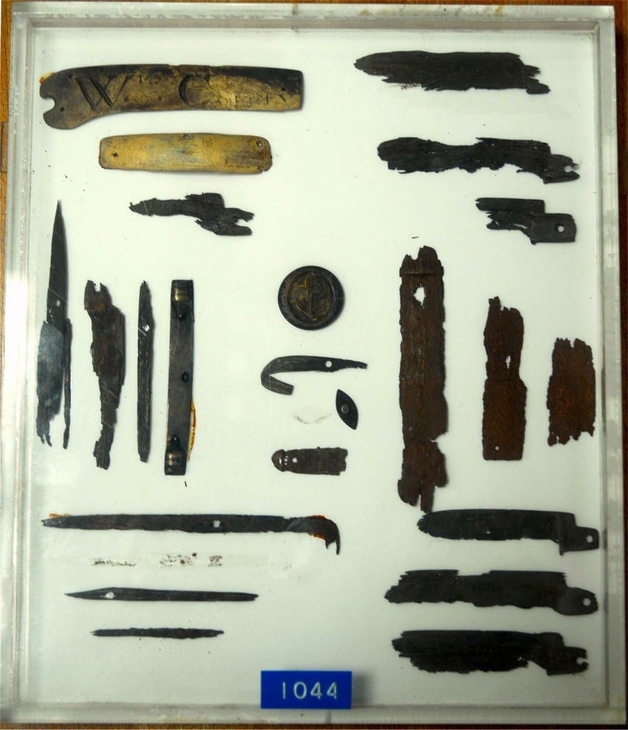 A modern photograph of a museum display case containing fragments of corroded knife blades and some wooden handles, one of which has the initials W C. 