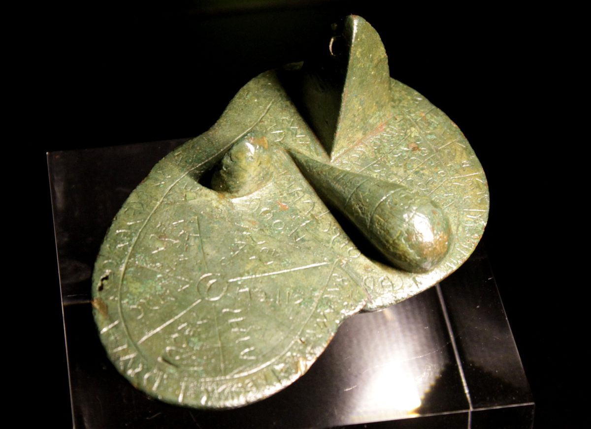 Modelo de bronze do fígado de um carneira, de coloração meio esverdeada, meio desbotada. Cheio de inscrições e com três relevos arredondados.