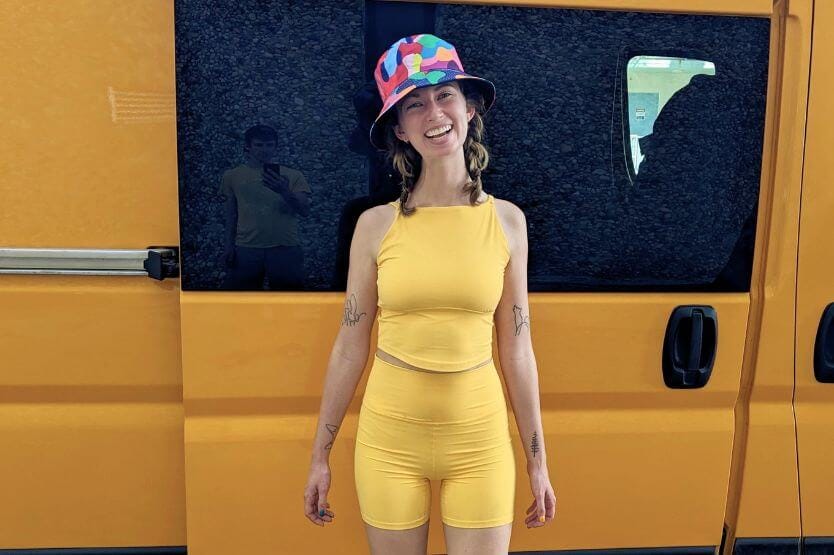 Haley, a young blonde woman, stands in front of the sliding door of a bright yellow camper van while wearing a yellow workout outfit and a brightly colored hat