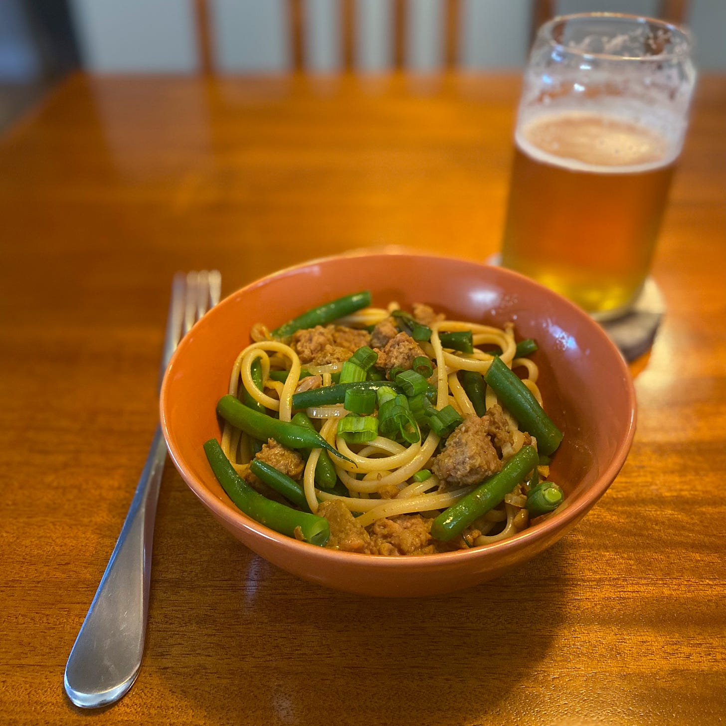 An orange bowl of the drunken noodles, with pieces of green beans and beyond meat throughout. Sliced green onions garnish the top, and a glass of beer sits on a coaster behind the bowl.