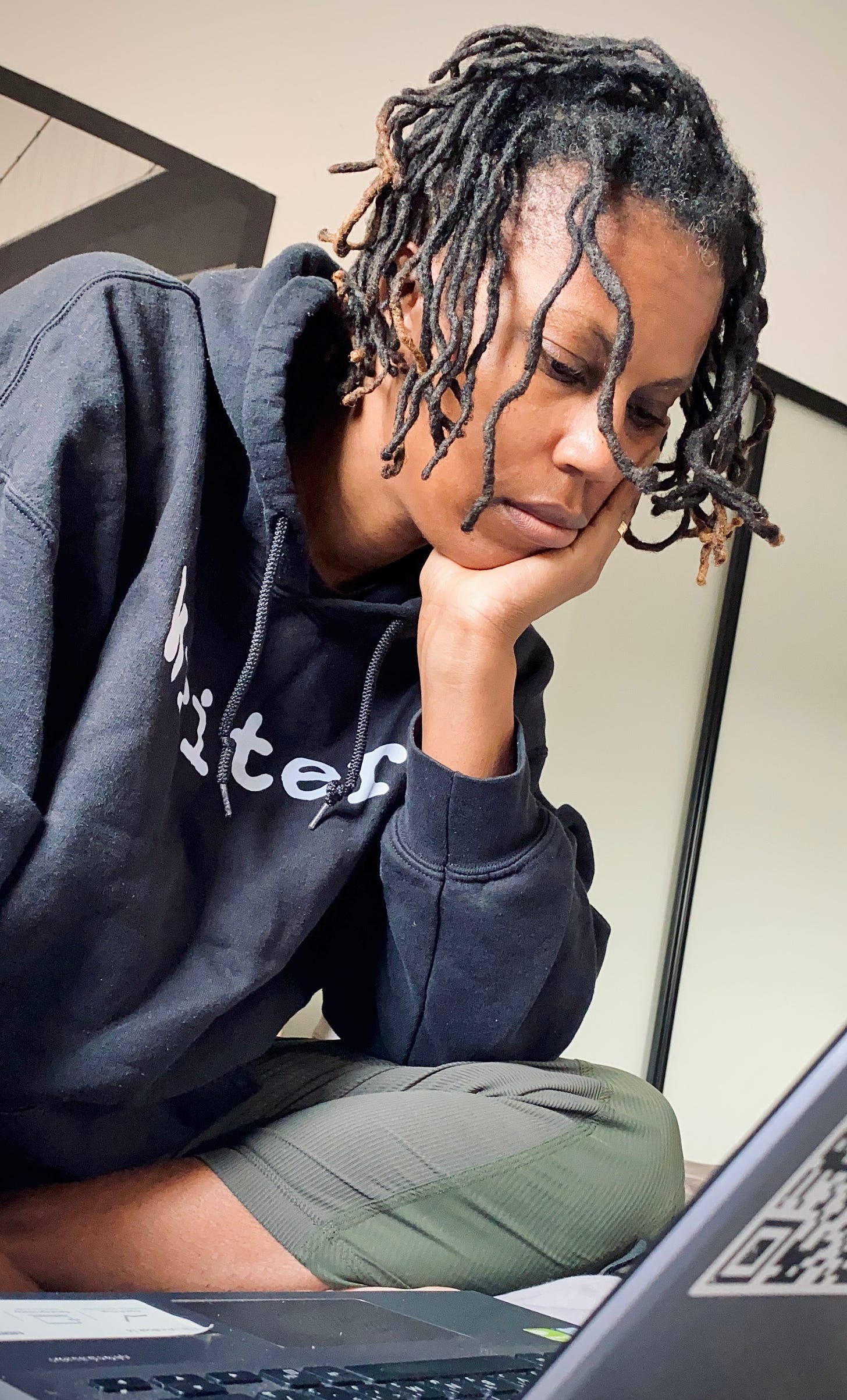 Sandra has black locs, wears a hoodie that says writer on it, sits in front of their laptop