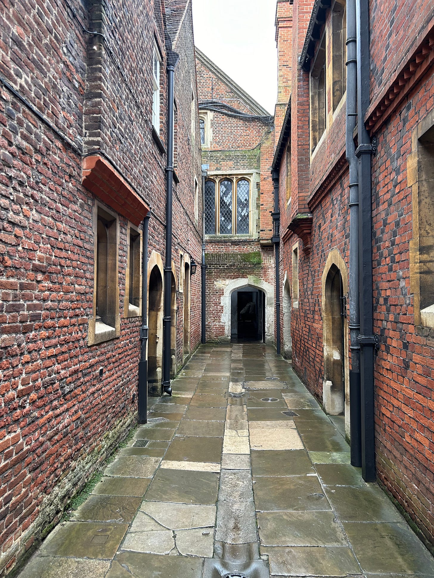 A red brick series of walls, passages, and windows, obviously old. It is a wet and grey day, and the flagstones of the alleyway are wet.