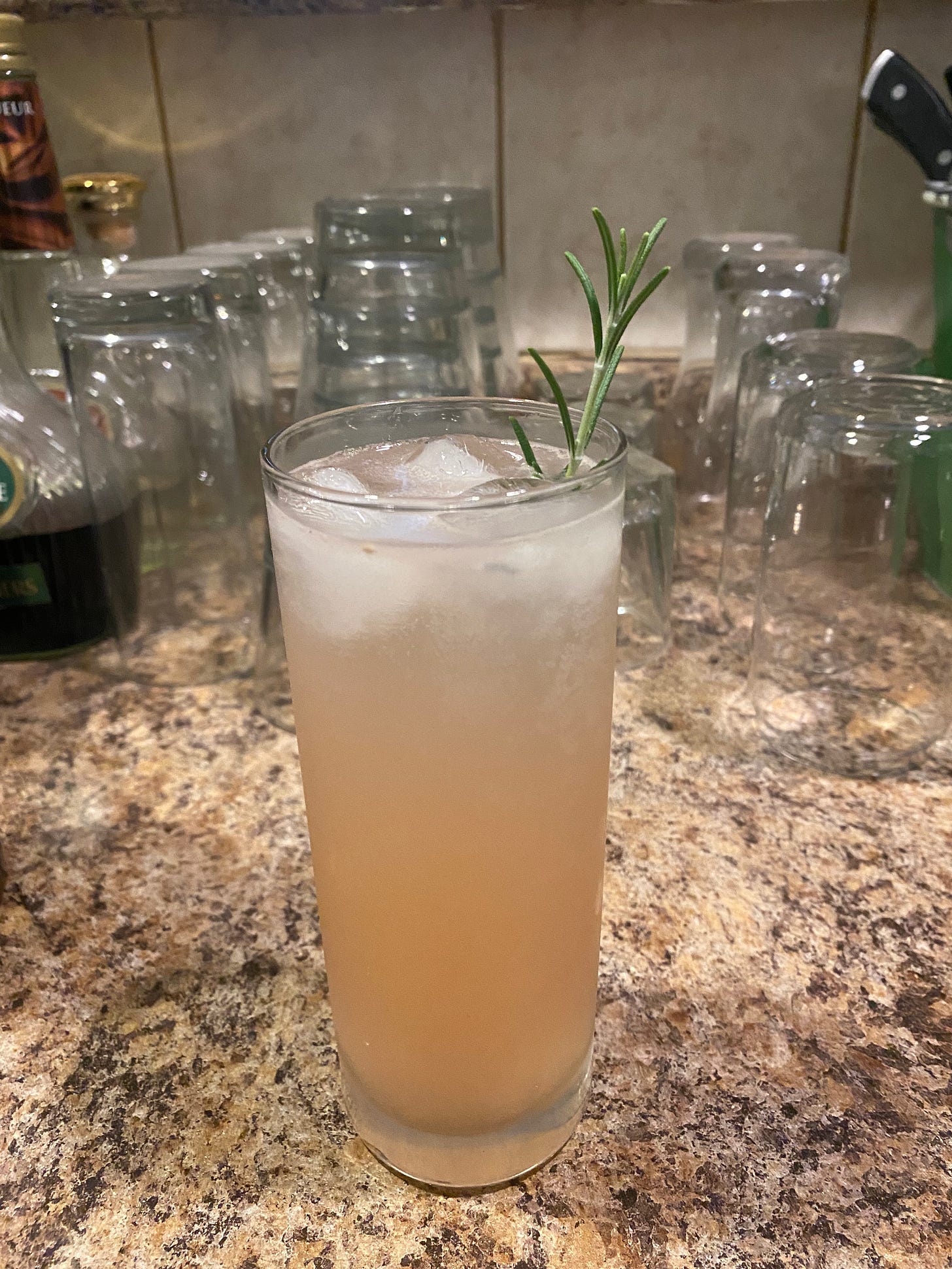 In a collins glass, the drink described below. Ice cubes float at the top, and a sprig of rosemary sticks out of the glass on the right. The drink is pale pink in colour. Behind it on the bar are many overturned empty glasses.