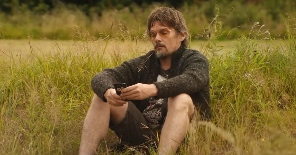 Still from Juliet Naked that shows Ethan Hawke sitting in the tall grass, a weed in his mouth, looking down at his phone as he emails back and forth with Annie.
