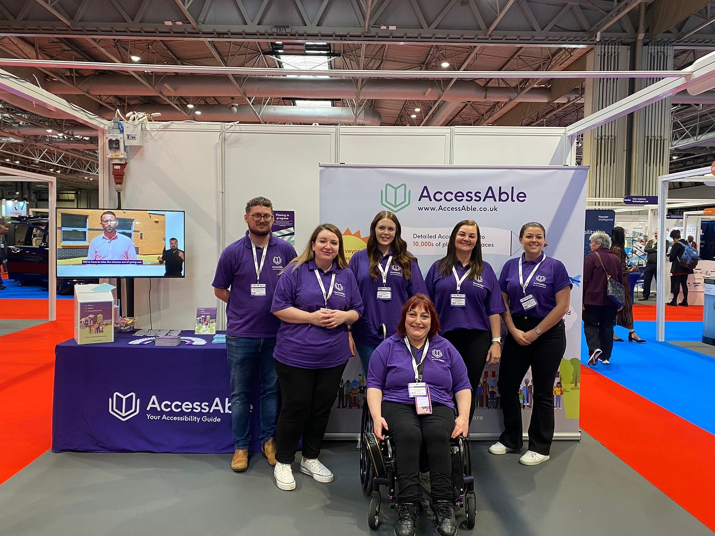 5 white women and 1 white man in purple shirts take a team photo at Naidex on the AccessAble stand. 