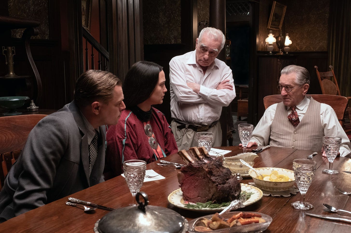 Leonardo DiCaprio, Lily Gladstone, and Robert De Niro in constume and character at a dinner table on the set of Killers of the Flower Moon while Director Martin Scorsese stands with them talking