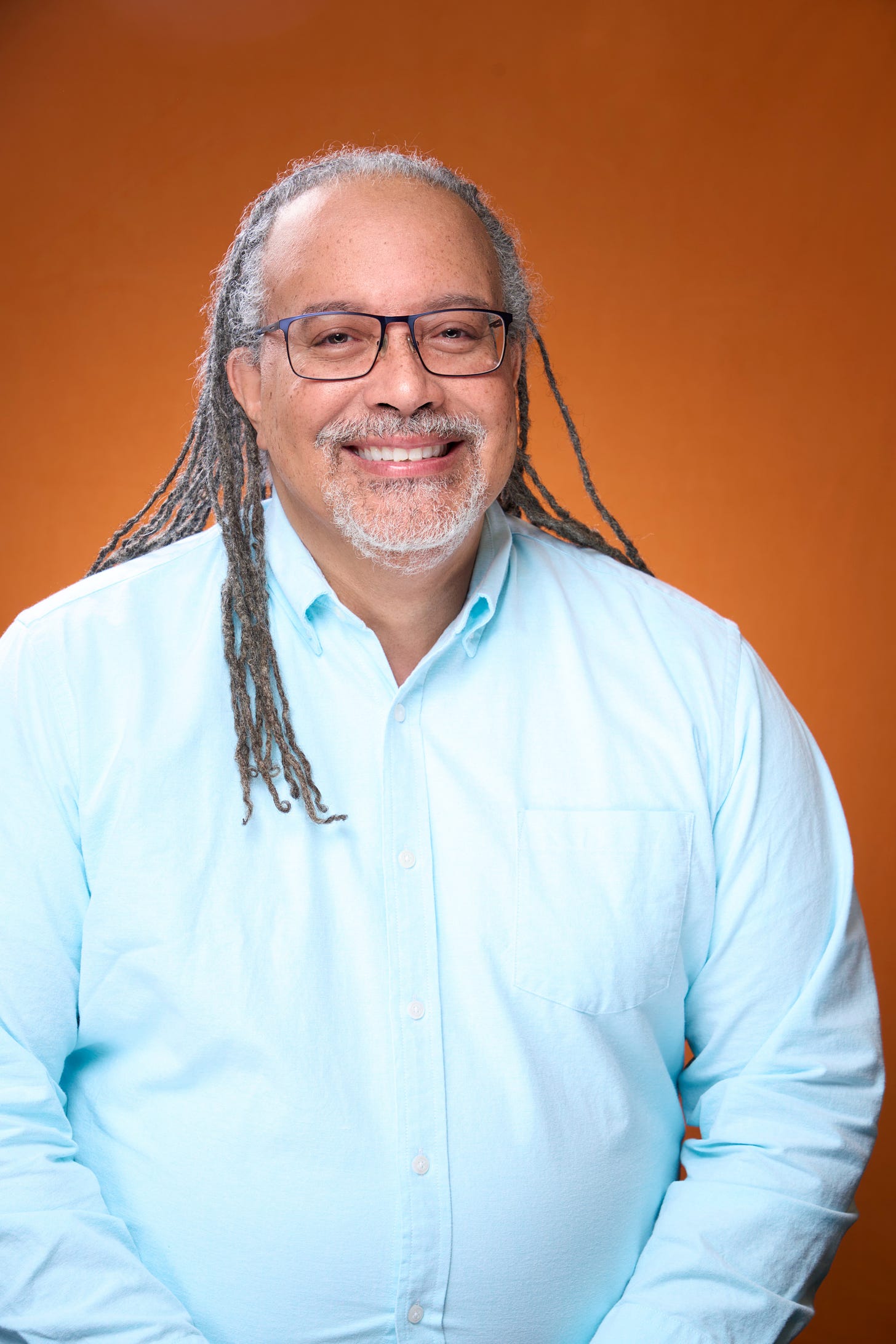 Lovely headshot photo of Ludovic Blain, in a blue collared button down shirt. Smiling, wearing glasses, with light brown skin and thin dreadlocks down to his chest.