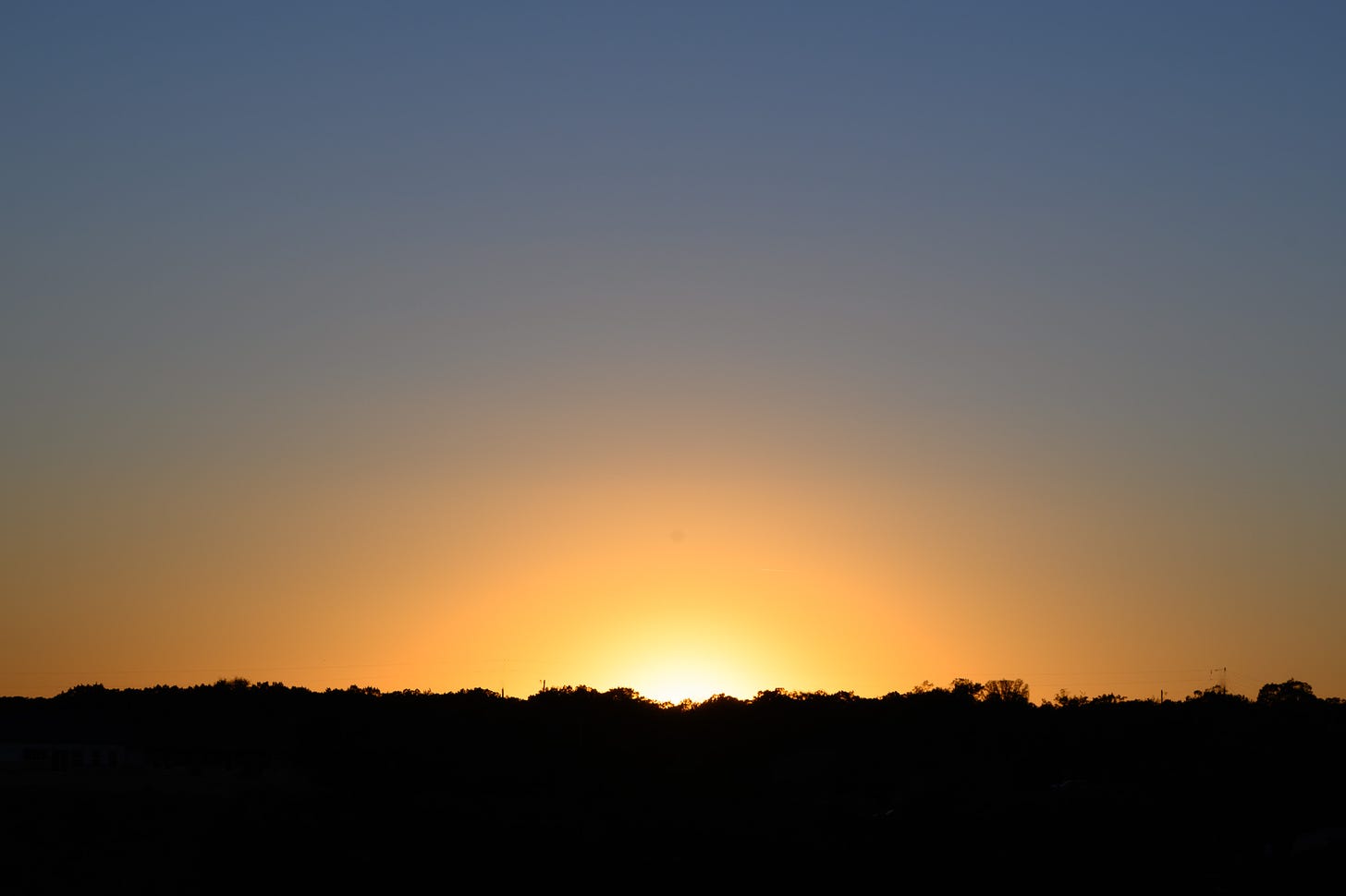 The setting sun above a treeline with a clear blue sky above and the sun’s rays forming a semi-circle above the horizon