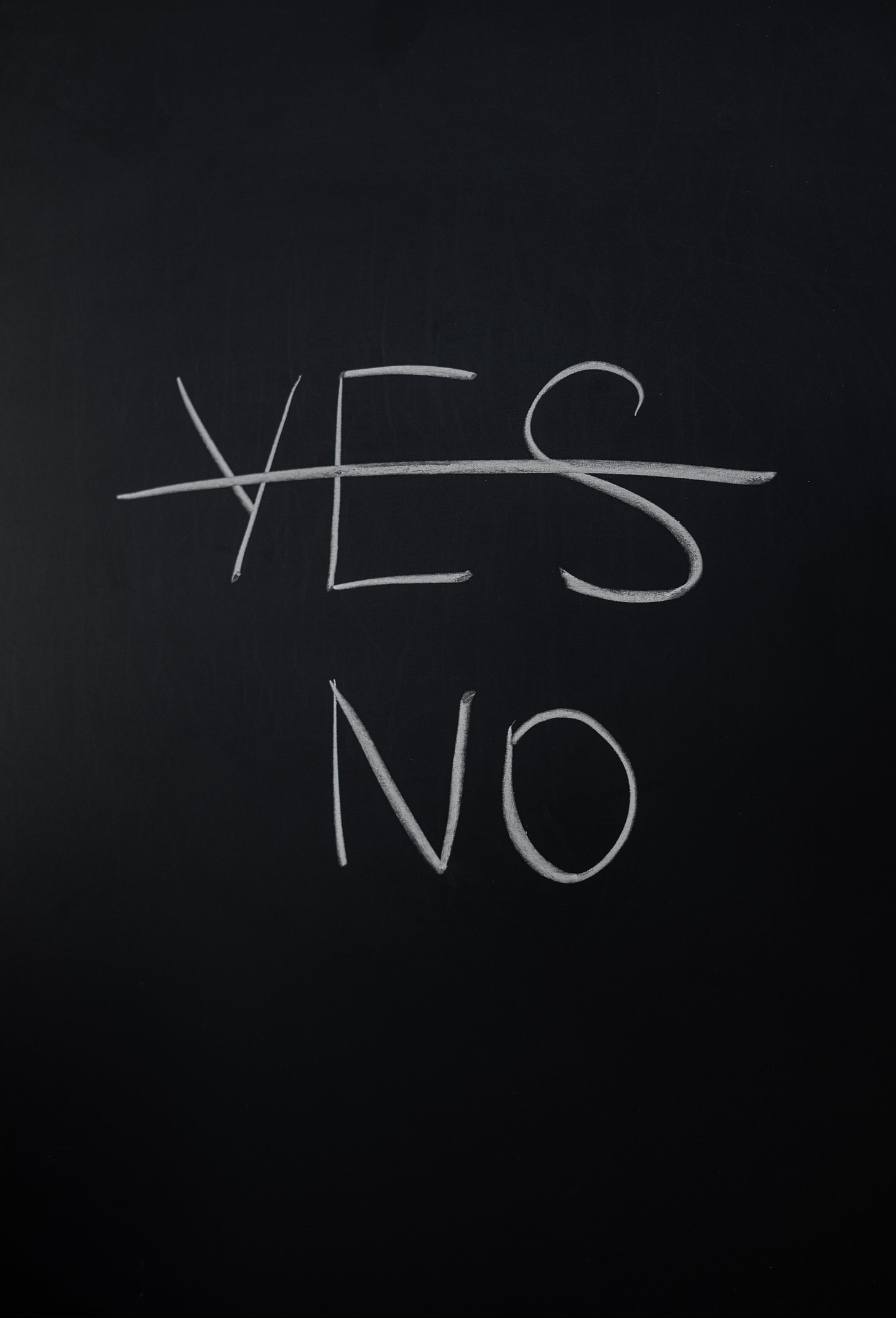 The word “yes” is crossed out on a chalk board in white chalk, leaving only the word “no” below it (also in white).