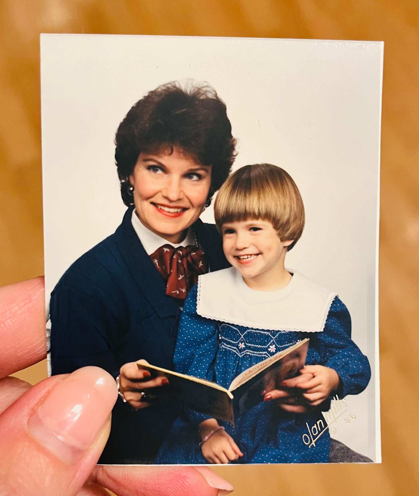 A woman with a little girl on her lap. The little girl is holding on side of an open book, the woman (mother) is holding the other