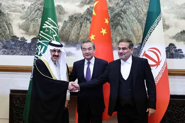 A Chinese official stands between two representatives from Iran and Saudi Arabia, who are shaking hands. Flags of the officials’ countries appear behind them.