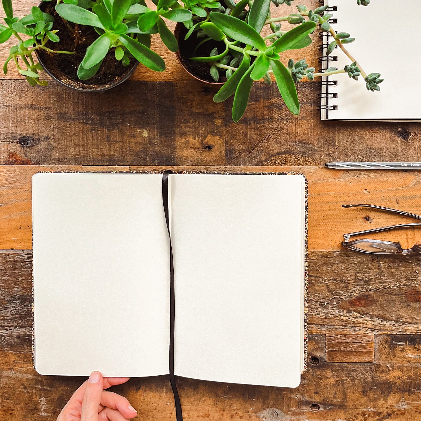 Caderno em branco aberto sobre uma mesa de madeira, com vasos de plantas, bloco de notas e óculos