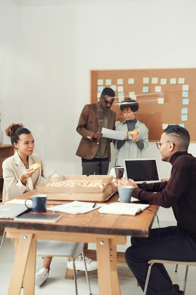 People sitting around a table eating pizza while looking at work or talking. There are notes scattered around, and two people talking while sitting at the table while two people are at the back looking over a sheet of paper