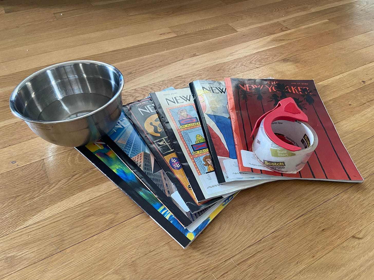 A metal bowl filled with water, a variety of The New Yorker magazines, and packaging tape laid out on a wooden floor.