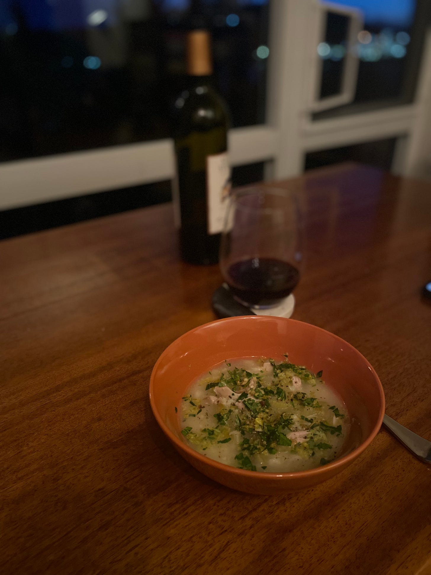 An orange bowl of the soup described above, with gremolata scattered overtop. In the background is a bottle of wine, and a glass of red wine on a coaster in front of the window.