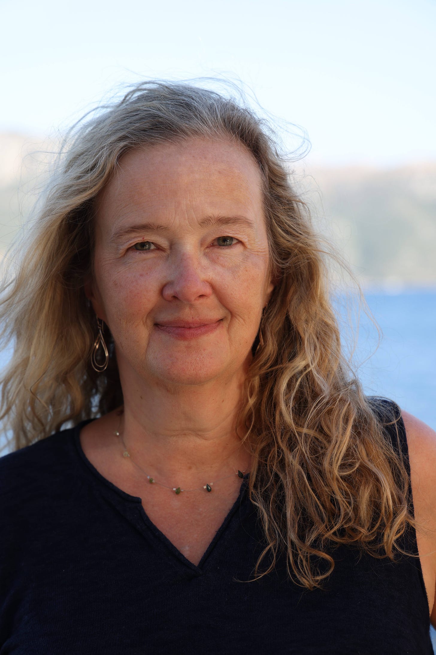 A woman stands on a beach, smiling serenely into the camera, her curly hair falling around her shoulders. 