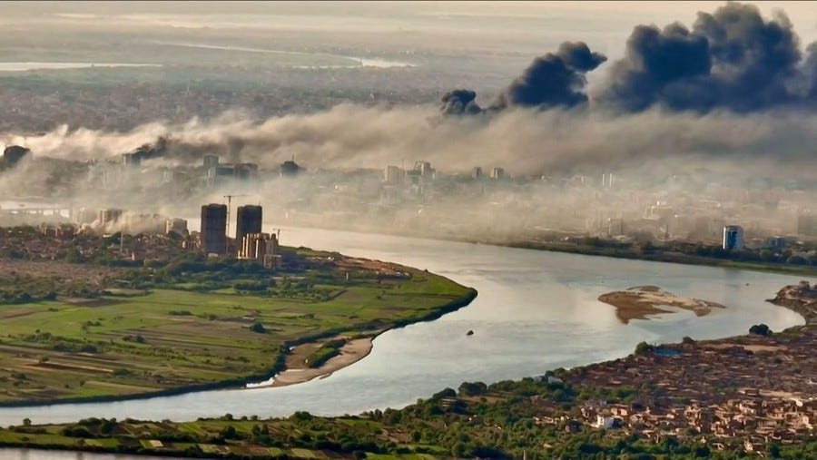 An elevated view of a river running through a city with black and white smoke rising from numerous fires.