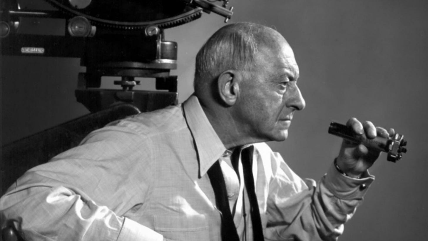 Director Cecil B. DeMille, in profile, holds a viewfinder and looks sternly out of shot - in the background a 35mm film camera