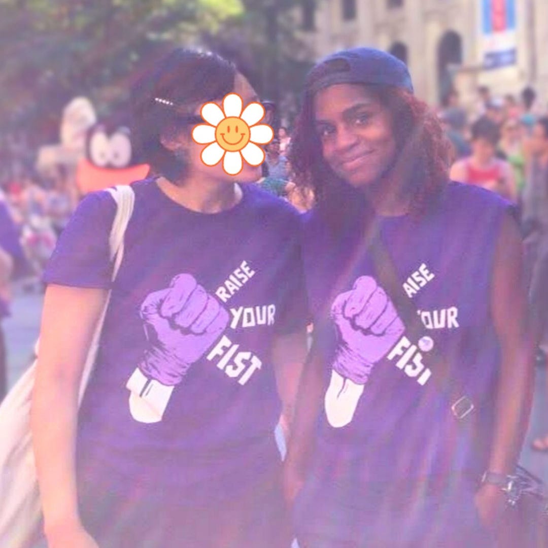 Jasper stands arm in arm with nondescript asian friend wearing 'raise your fist' dyke march shirts