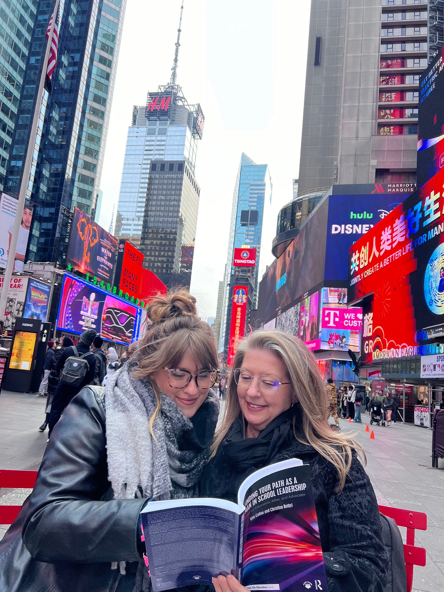 The author and her daughter seated on red folding chairs reading the book, Finding Your Path as a Woman in School Leaderhsip. They are seated in the vibrant setting of Times Square in New York City.