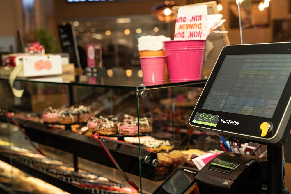 A two-level counter and display with sprinkled donuts, bright pink cups, and other gift items from Dunkin along with a cash register
