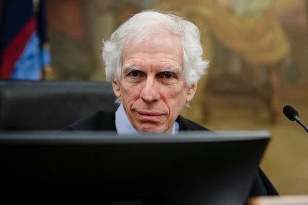 Arthur Engoron, in a blue shirt under a black judicial robe, looks directly at the camera over a computer monitor on his bench in the New York State Supreme Court.