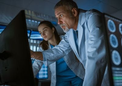 a middle-aged male scientist wearing a white lab coat points at a computer screen while a younger woman scientist also wearing a lab coat looks on.