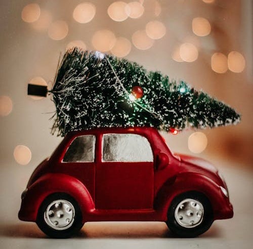 Free A red toy car carries a Christmas tree, set against a warm bokeh background, celebrating the festive season. Stock Photo