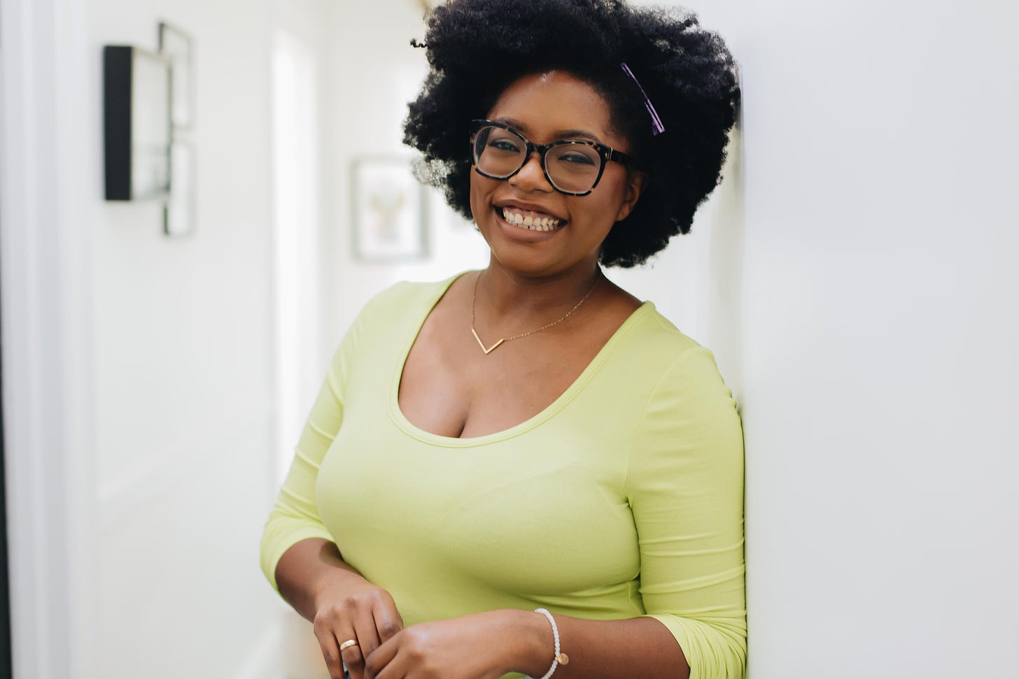 Black woman with a fro and glasses wearing a long-sleeve lime green shirt and a gold L initial necklace standing in a long white hallway