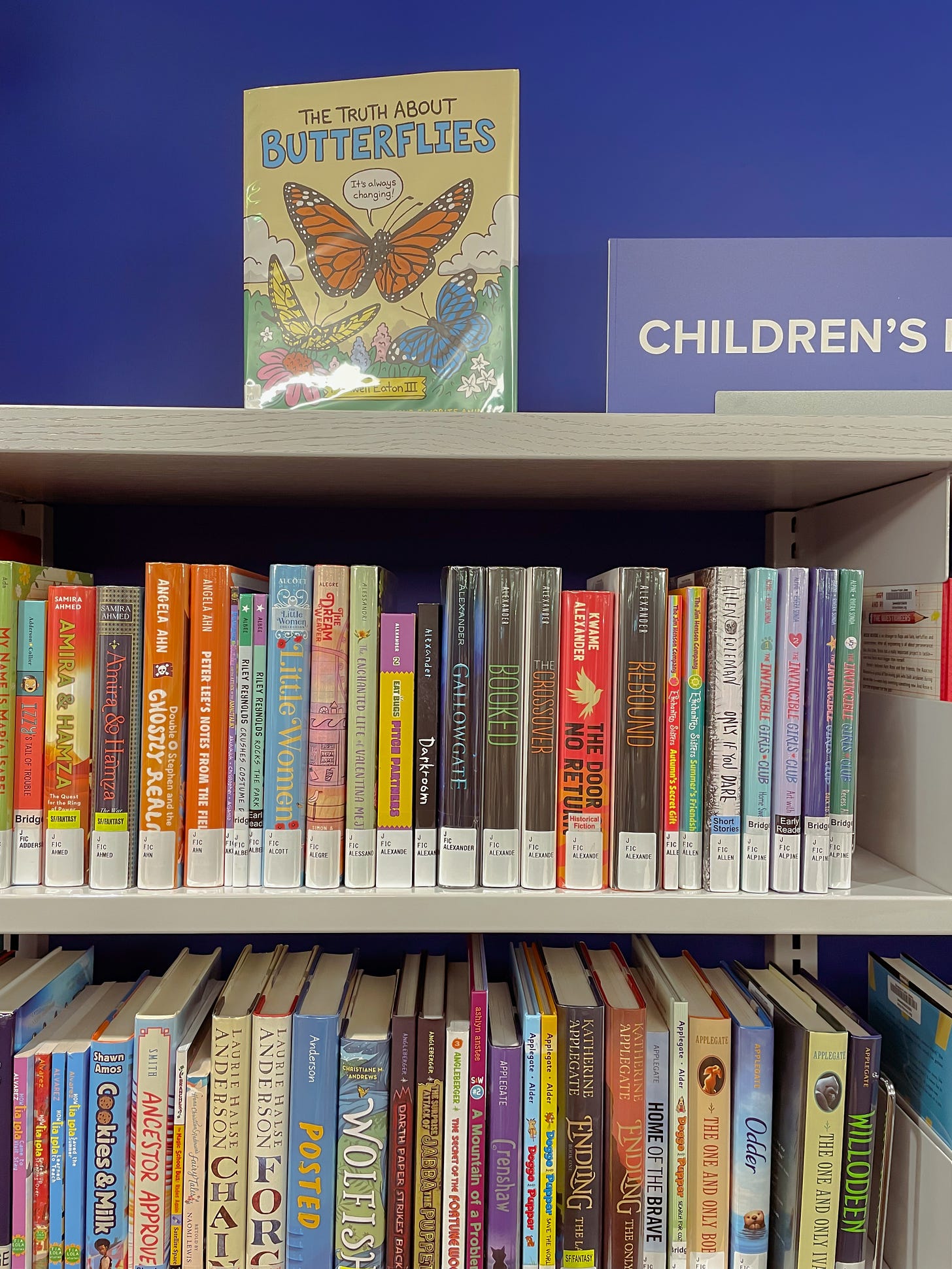 Another photo of the children's and middle grade books shelf at a public library. the wall behind is dark blue. a sign on top of the shelf reads: children's books.