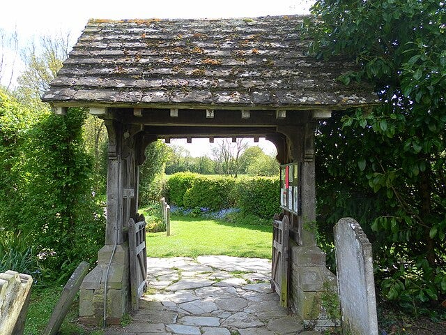 A lychgate with a tiled roof and stone walls. The wooden gates stand open, revealing a green lawn beyond. A lychgate with a tiled roof and stone walls. The wooden gates stand open, revealing a green lawn beyond.