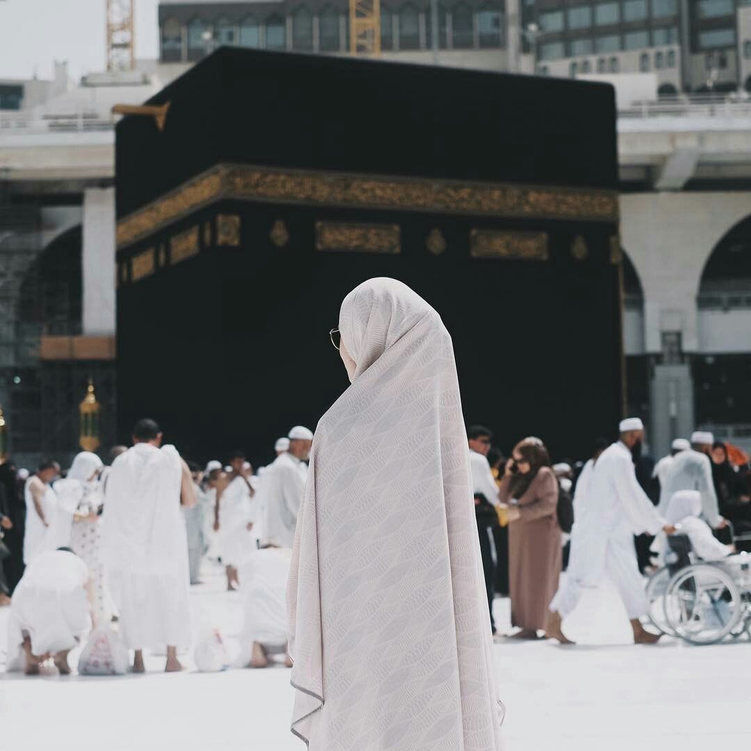 Woman in white jilbab and glasses faces left, edge of face barely visible. She is standing some distance away from the black and gold Ka'aba, and other Hajj pilgrims clad in white are between her and the Ka'aba.