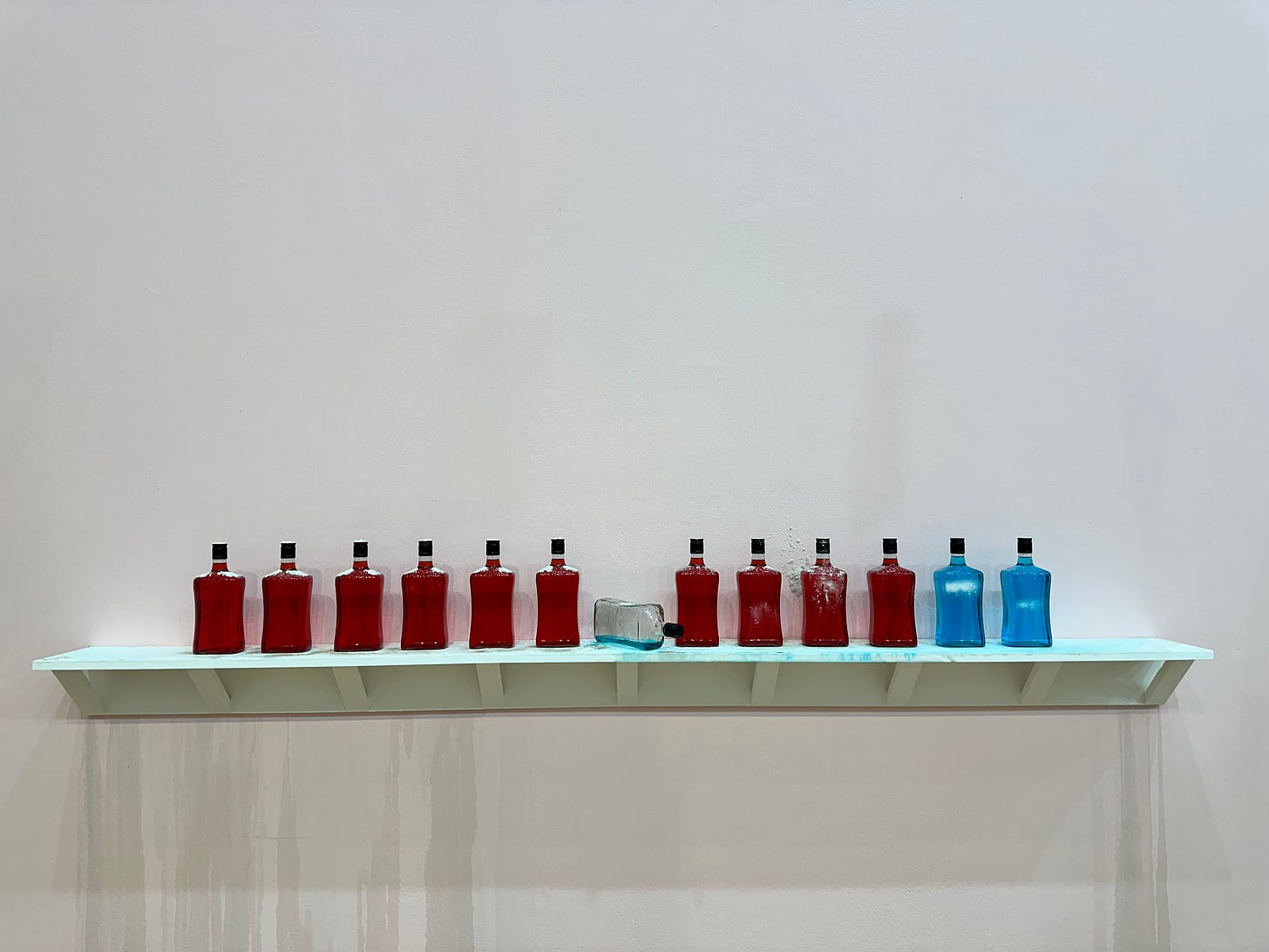 A line of 13 red and turquoise glass bottles on a white ledge, in front of a white wall, where one bottle has tipped over and its liquid has run down the wall.