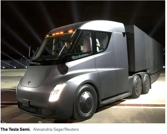 A Tesla Semi truck is on a test drive, metal gray with lights on and a driver turning the truck